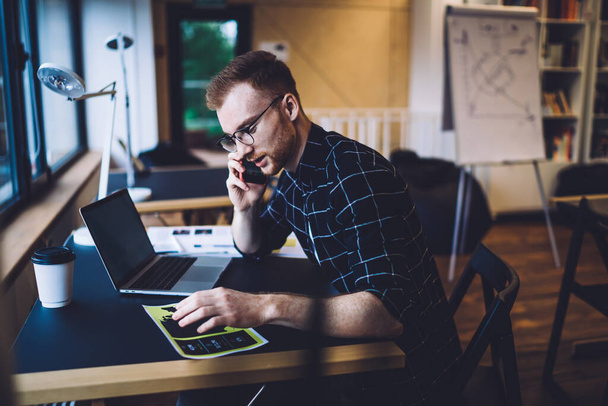 Caucasian male programmer sitting at table desktop and using mobility connection for making cellphone communication and discussing database details, smart casual copywriter phoning via gadget - 写真・画像