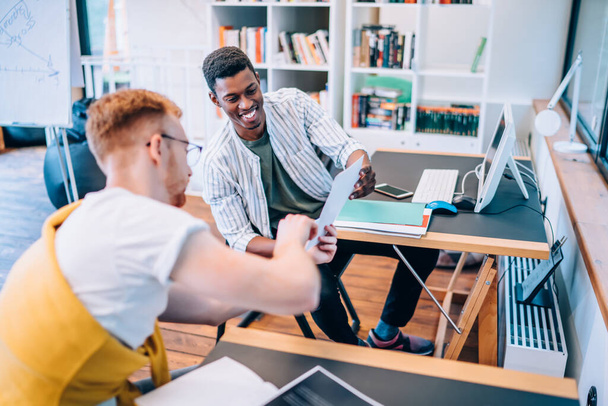 Young guy in casual clothes showing paper with data to cheerful African American coworker while sitting at tables and working at startup project together - 写真・画像