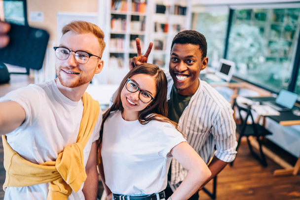 Delighted African American guy in casual clothes smiling and showing V sign while taking selfie with positive young colleagues in contemporary office - Zdjęcie, obraz