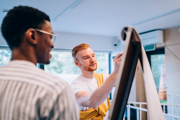Young male drawing scheme on flip chart while standing near interested African American coworker while working on creative project in modern office - Foto, Bild