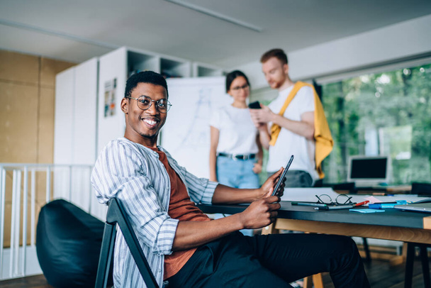 Cheerful smiling smart diverse young coworkers in casual apparel surfing smartphone watching tablet  in office space with bookshelf opposite of big window   - Foto, Imagen