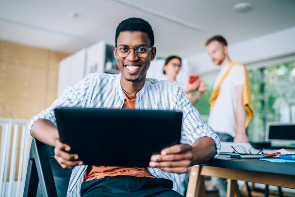 From below of joyful smiling African American man in casual wear and glasses using tablet messaging with friend looking at camera in office - Foto, Imagen