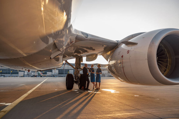 Letadlo poblíž terminálu na letišti při západu slunce. Pilot a dvě letušky stojí spolu a mluví po přistání nebo před odletem - Fotografie, Obrázek