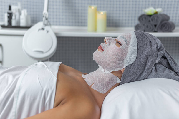 Vista lateral de uma mulher latina relaxante durante o tratamento de cuidados com a pele com gazes no rosto no salão de spa. Conceito de cuidados da pele. - Foto, Imagem
