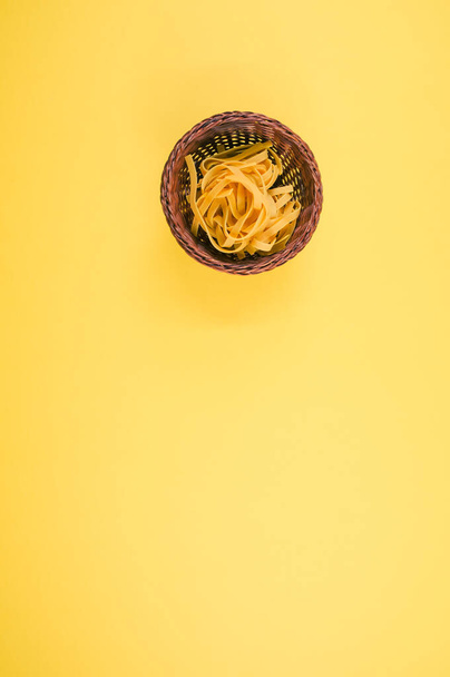 A vertical shot of uncooked fettuccine pasta on a wicker bowl isolated on yellow background - Фото, изображение
