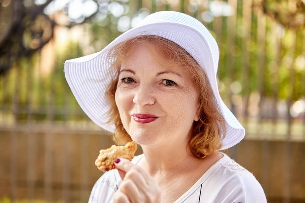 Smiling aged white woman eats cookie outdoors. - 写真・画像