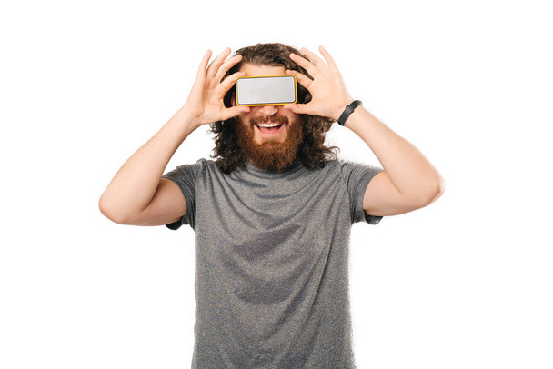 Smiling bearded man is covering his eyes with smart phone over white background. - Photo, Image