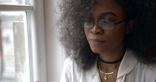 Young attractive afro-american woman in glasses is drinking tea while sitting on the window sill. Close-up portrait. 4k. - Filmati, video