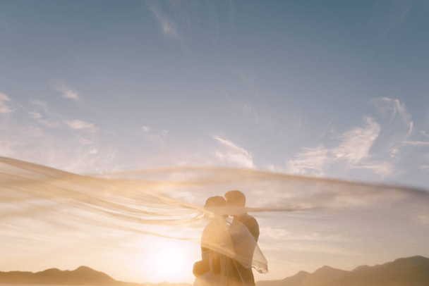 A vőlegény megcsókolja a menyasszonyt a lebegő fátyol alatt a hegyek és az ég hátterében. - Fotó, kép