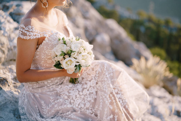 Bruid met een boeket pioenen zit op de rotsen tegen de achtergrond van bergen en groen - Foto, afbeelding