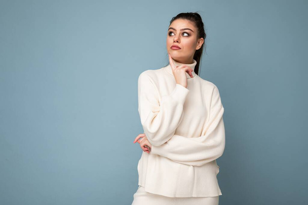 Young thoughtful brunette woman with wearing white casual sweater isolated over blue background looking confident at the camera with crossed arms and hand raised on chin. Thinking positive - Photo, Image