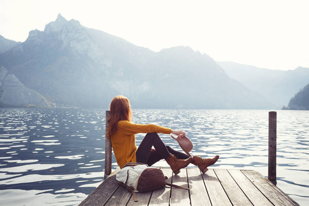 beautiful girl in a hat sitting on the pier at the lake bank and mountains in the backgroun - Foto, imagen