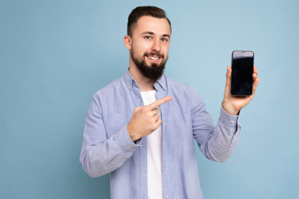 Przystojny szczęśliwy, fajny młody człowiek dobrze wyglądający, noszący stylowe ubrania stojący w izolacji nad kolorową ścianą tła, trzymający smartfona i pokazujący telefon z pustym ekranem, patrzący na - Zdjęcie, obraz