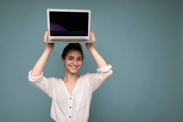 Hübsch lächelnde junge Frau hält Netbook-Computer in die Kamera und trägt weißes Hemd isoliert auf blauem Hintergrund - Foto, Bild