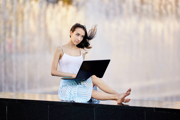 Slender woman with laptop sits near fountain in city garden in summer. - Photo, Image