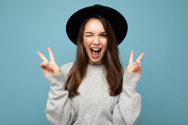 Photo of young positive happy adorable attractive pretty brunette woman with sincere emotions wearing casual grey sweater and black hat isolated over blue background with free space and showing peace - Photo, Image