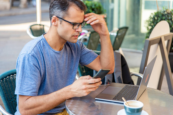 Jonge Latijnse man gestrest werken met mobiele telefoon en computer buiten. - Foto, afbeelding