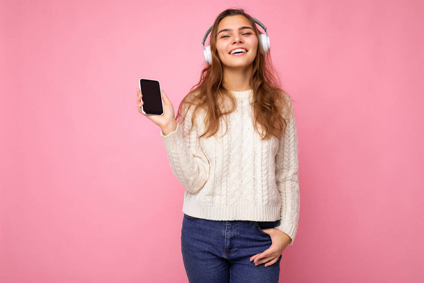 Foto de bela jovem mulher vestindo suéter elegante branco claro isolado no fundo rosa vestindo fones de ouvido sem fio Bluetooth branco e ouvir música legal e mostrando telefone com vazio - Foto, Imagem