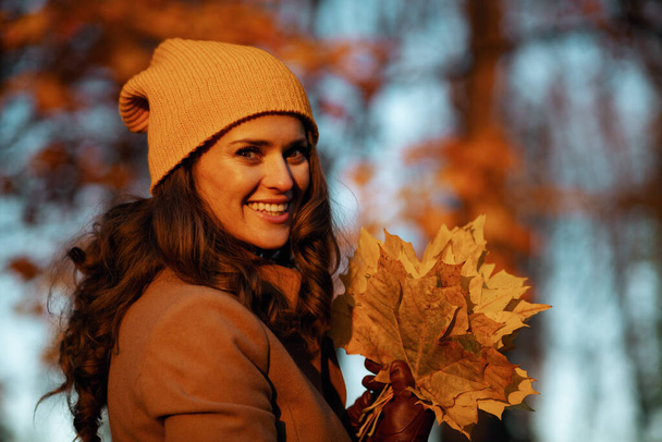 Hello october. smiling elegant middle aged woman in brown coat and yellow hat with autumn yellow leaves outside in the city park in autumn. - Fotó, kép