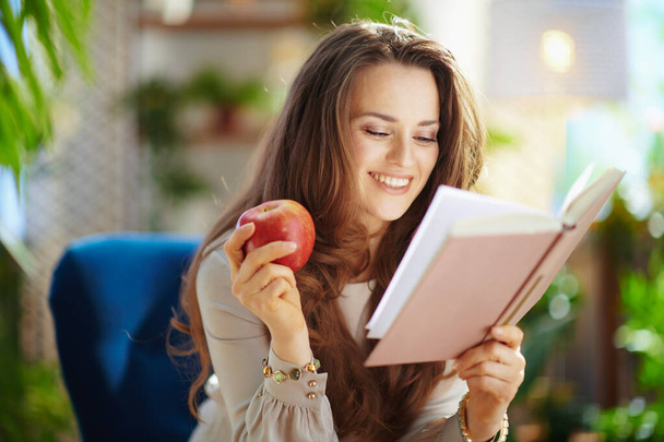 Green Home. happy stylish woman with long wavy hair with book and red apple sitting in a blue armchair in the modern house in sunny day. - Foto, Bild