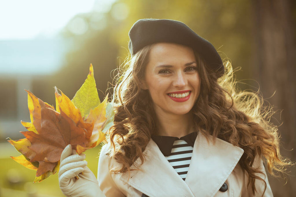 Hello september. Portrait of happy modern woman in beige trench coat and black beret with autumn yellow leaves outdoors in the city in autumn. - Foto, immagini