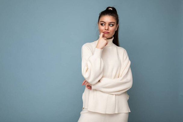 Young brunette woman with wearing white casual sweater isolated over blue background looking confident at the camera with crossed arms and hand raised on chin. Thinking positive - Фото, зображення