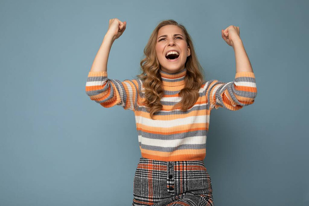 Portrét emocionální šťastný pozitivní mladé atraktivní blondýny kudrnaté ženy s upřímnými emocemi na sobě trendy pruhované pulovr izolované na modrém pozadí s kopírovacím prostorem a slaví vítězství - Fotografie, Obrázek