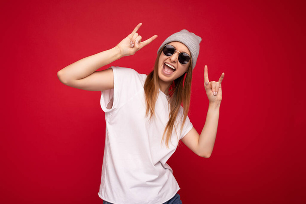 Photo shot of beautiful positive young dark blonde woman wearing casual clothes and stylish sunglasses isolated over colorful background wall looking at camera having fun and showing rock and roll - Foto, afbeelding