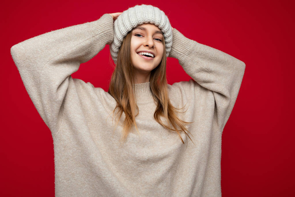 Shot of attractive positive joyful amusing young dark blonde female person standing isolated over red background wall wearing beige warm sweater and winter beige hat looking at camera - Photo, Image