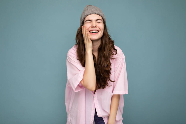 Güzel, pozitif, duygusal, esmer bir kadının mavi arka plan duvarında dikilip pembe gömlekli, gri şapkalı ve kahkahalarla güldüğü bir fotoğraf. - Fotoğraf, Görsel