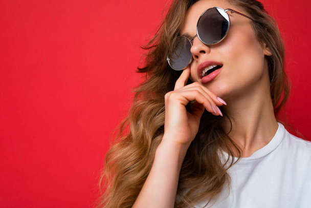 Closeup shot of beautiful sexy thoughful young dark blonde curly woman isolated over red background wall wearing casual white t-shirt and stylish sunglasses looking up and thinking - Photo, Image