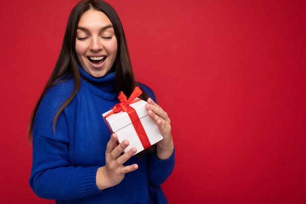 Foto de una joven morena sorprendida bastante positiva aislada sobre una colorida pared de fondo con un atuendo de moda que sostiene la caja de regalo y mira la caja actual con cinta roja - Foto, Imagen