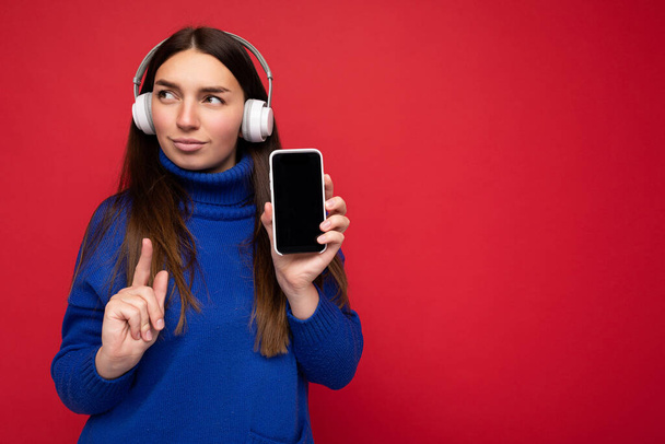 Vrij ernstige geconcentreerde jonge brunette vrouw dragen blauwe trui geïsoleerd over rode achtergrond houden en tonen mobiele telefoon dragen witte oortjes luisteren naar muziek kijken naar de zijkant en - Foto, afbeelding