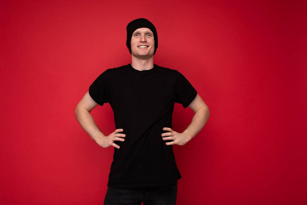 Photo d'un beau jeune homme souriant et heureux debout isolé sur un mur de fond rouge portant un t-shirt noir pour la maquette et le chapeau noir et regardant la caméra - Photo, image