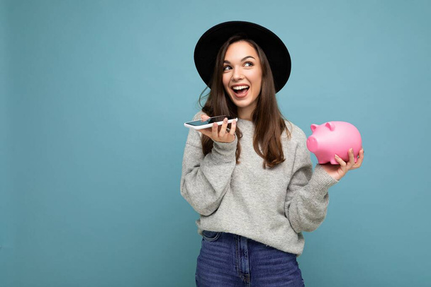 Happy positive smiling laughing young attractive brunette woman wearing trendy grey pullover and black hat isolated over blue background with free space, holds pink piggy bank and uses smartphone - Photo, Image