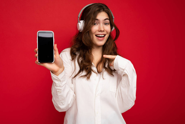 Красивая счастливая улыбающаяся молодая женщина в стильной повседневной одежде, изолированной на фоне стены, держа и показывая мобильный телефон с пустым дисплеем для макета в белых наушниках Bluetooth - Фото, изображение