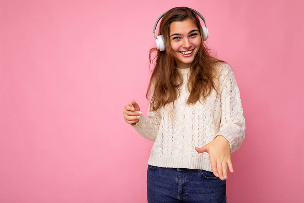 Красива щаслива усміхнена молода брюнетка кучерява жінка в білому светрі ізольована на рожевій стіні в білих навушниках Bluetooth, слухаючи прохолодну музику, дивлячись на камеру
 - Фото, зображення