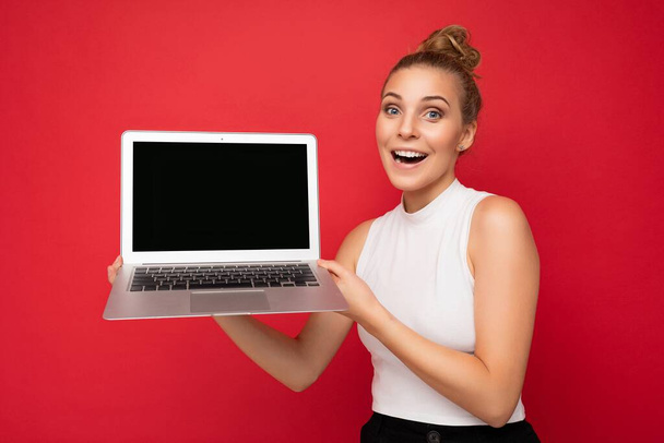 χαμογελαστή χαρούμενη όμορφη ξανθιά νεαρή κοπέλα με μαζεμένα μαλλιά κοιτάζοντας κάμερα κρατώντας φορητό υπολογιστή με άδεια οθόνη οθόνης με mock up και αντίγραφο χώρου φορώντας λευκό t-shirt που απομονώνονται στον κόκκινο τοίχο - Φωτογραφία, εικόνα