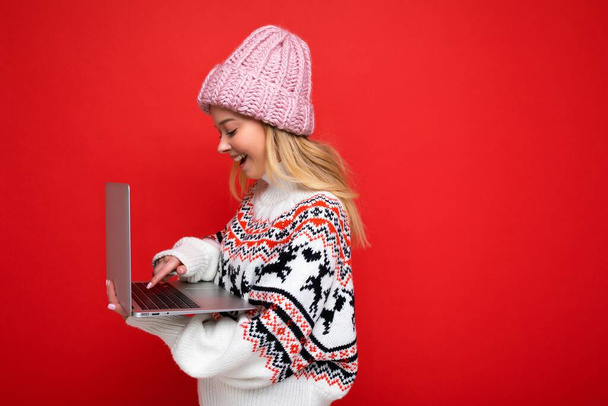 side profile photo of charming smiling happy pretty young lady holding laptop isolated over red wall background looking at monitor screen wearing winter hat and knitted new year sweater - Φωτογραφία, εικόνα