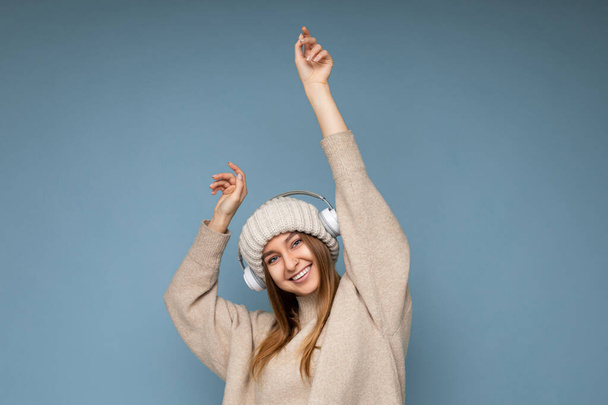 Привлекательная позитивная улыбающаяся молодая блондинка в бежевом зимнем свитере и шляпе на синем фоне стены в белых беспроводных наушниках Bluetooth слушая музыку, танцующую весело - Фото, изображение