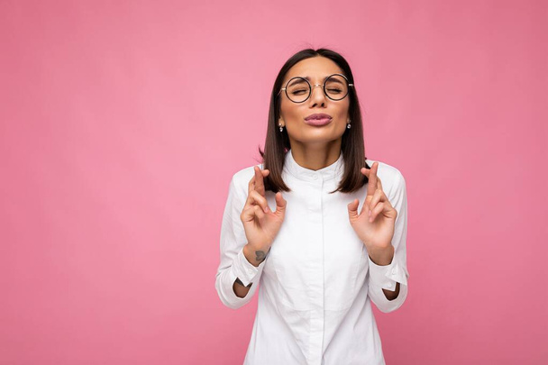 Portrait of young emotional pretty brunette lady with short haircut wearing casual white shirt and optical glasses isolated over pink background with copy space and crossing fingers and hoping for - Photo, Image