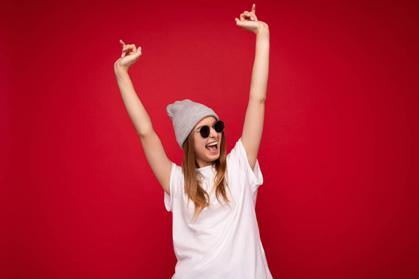 Photo shot of beautiful positive smile young dark blonde woman isolated over red background φορώντας γκρι καπέλο casual λευκό t-shirt για mockup και κομψά γυαλιά ηλίου στο πλάι και διασκεδάζοντας - Φωτογραφία, εικόνα
