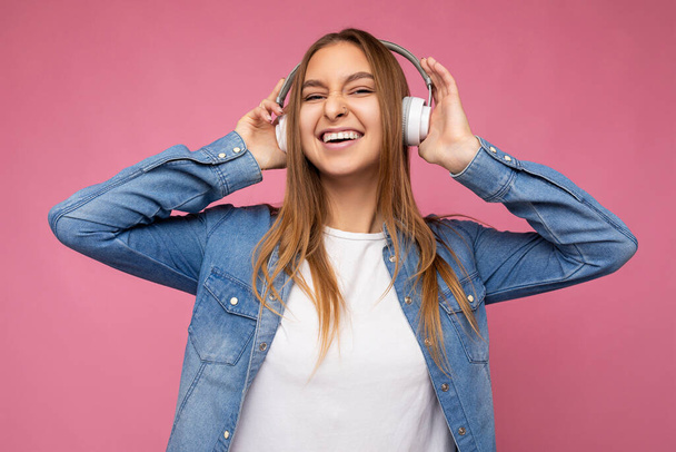 Photo of beautiful happy smiling young blonde woman wearing blue jean shirt and white t-shirt isolated over pink background wearing white wireless bluetooth earphones listening to cool music and - 写真・画像