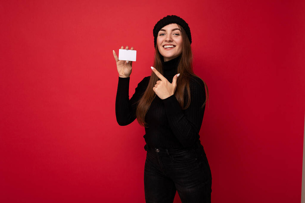 Foto de bela jovem morena vestindo suéter preto e chapéu isolado no fundo vermelho segurando cartão de crédito olhando para a câmera e apontando o dedo para o cartão de plástico - Foto, Imagem