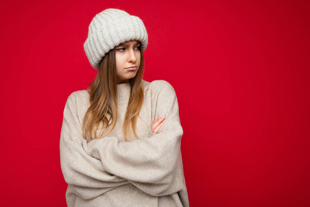 Photo shot of beautiful offended resentful young dark blonde woman standing isolated over red background wall wearing warm sweater and winter hat looking to the side with crossing hands - Foto, Imagem
