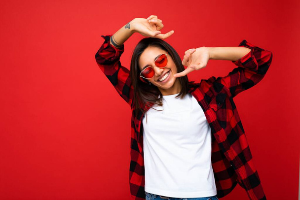 Portrét pozitivní veselý úsměv mladé brunetky žena v ležérní bílé tričko pro mockup, stylové červené kontrolní tričko a trendy červené sluneční brýle izolované na červeném pozadí s kopírovacím prostorem - Fotografie, Obrázek