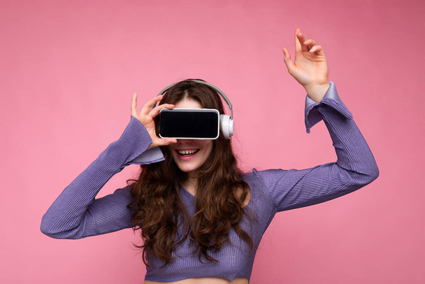 Photo of beautiful happy young brunette curly woman wearing purple crop top isolated over pink background wall holding and suggesting mobile phone with empty display and bluetooth earphones - Fotó, kép