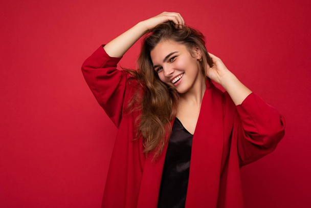 Foto portret van een mooie vrolijke lachende jonge brunet vrouw in stijlvol rood vest. Sexy zorgeloze vrouwelijke persoon poseren geïsoleerd op rode achtergrond. Positief model toont oprechte emoties - Foto, afbeelding