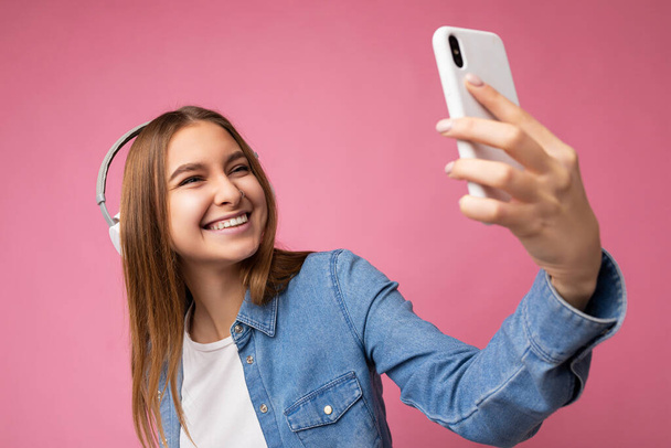 Foto van mooie vrolijke glimlachende jonge blonde vrouw met blauw jean shirt en wit t-shirt geïsoleerd over roze achtergrond met witte bluetooth hoofdtelefoon en het luisteren naar muziek en het gebruik van mobiele - Foto, afbeelding