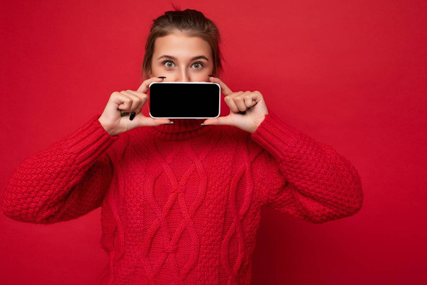 Photo of beautiful young woman wearing warm red sweater isolated over red background wall holding smartphone and showing phone with empty display for mockup looking at camera - Fotó, kép
